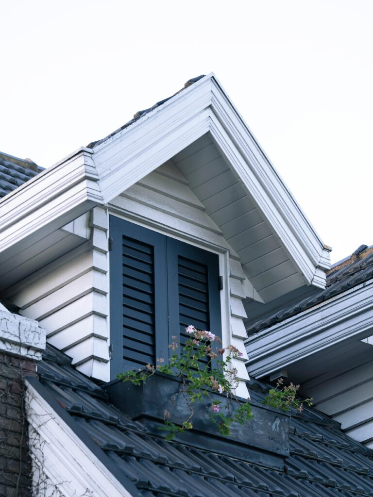 Gable roof with a dormer window featuring shutters and a flower box against a clear sky backdrop.
